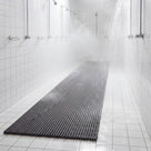 Dark grey Heronrib matting running the length of a white-tiled communal shower block, shower water draining straight through the ribs onto the tiled floor beneath.