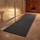 Dark grey Heronrib matting laid as a wet-side walkway across spa flooring between a glass-fronted steam room and a timber sauna door, draining into a stainless floor channel.