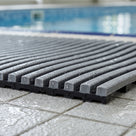 Close macro detail of dark grey Heronrib matting on a wet poolside tile, showing the finely ribbed top bars, the black channelled underbars beneath and the 10.5mm cut edge, with water draining through to the tile.