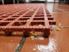 Close-up of the red open grid matting showing ribbed bars and debris dropping through to the tile beneath