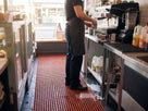 Red open grid matting behind a cafe counter with spilled milk draining through to the tiled floor
