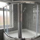 Chrome-framed curved quadrant shower enclosure over a bath, water-spotted glass doors closed, grey tiles behind