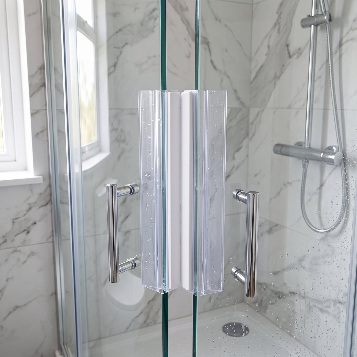 Two clear seals meeting down the edge of wet glass shower doors, white housing strip visible, marble tiles behind