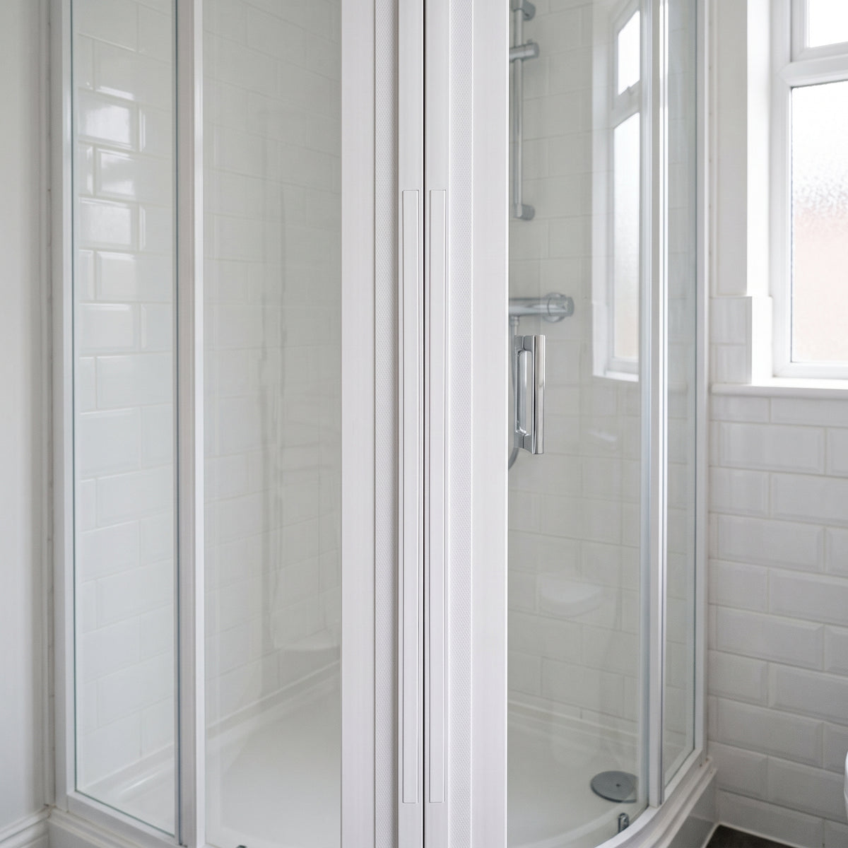 White-framed quadrant enclosure with curved sliding doors closed, white metro tiles and a window right