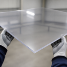Gloved hands holding a thick clear polycarbonate sheet by its corners, edge thickness visible, in a workshop