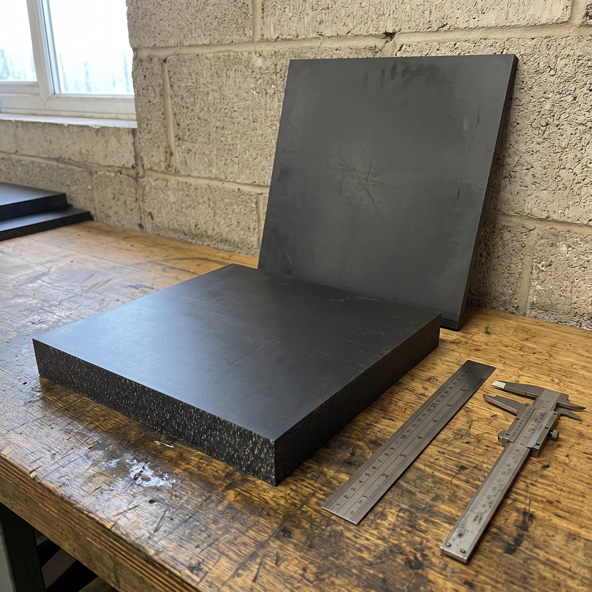 Thick black-grey nylon 6 slab on a workshop bench with a thinner slab leaning behind it, steel rule and callipers alongside