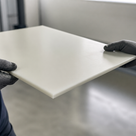 Slim ivory cast nylon panel supported at both edges by black gloves, grey workshop out of focus