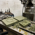 Thick green oil-filled cast nylon slabs on a milling machine table beside a milled cam plate cut from one, green swarf around