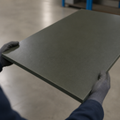 Grey gloves holding a rectangular olive-grey Oilon panel flat in a warehouse, blue racking behind