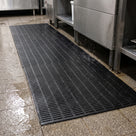 Black open-grid PVC matting unrolled along a commercial kitchen wash-up area beside a stainless steel sink, water draining through it onto the wet tiled floor.