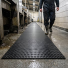 Black open-grid matting as a walkway through a wet industrial wash-down bay, worker in boots walking along it