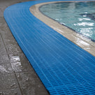 Blue open-grid matting laid as a continuous runner along the wet tiled surround of an indoor swimming pool, following the curve of the pool edge.