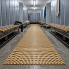 Beige open-grid PVC matting rolled out as a continuous runner down a changing room aisle between metal lockers and wooden benches.