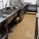 Beige open-grid matting along a commercial kitchen wash-up aisle, spare roll stored under the stainless sink
