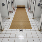 Beige open-grid matting unrolled between two rows of shower bays on white tiles, floor drain in the foreground