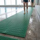 Green open-grid matting as a barefoot walkway on a wet leisure centre poolside, two swimmers walking along it