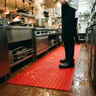 Red open-grid matting along a commercial kitchen hot line, chef at the range, spills draining through to the tiles