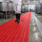 Red open-grid matting as a safety walkway through a wet food processing hall, worker in rubber boots, drain alongside