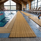 Beige open-grid matting running the length of a wet tiled indoor pool surround, swimmers in the lane beyond