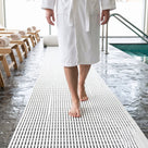 White open-grid matting forming a barefoot walkway across dark slate flooring in a hotel wellness area, running past loungers towards the pool edge.