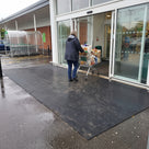 Black fine-ribbed open-grid entrance matting at a supermarket doorway, shopper pushing a loaded trolley in from a wet car park