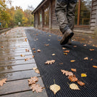 Black fine-ribbed open-grid matting strip on wet timber decking with autumn leaves, walker in boots stepping onto it