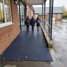 Oxford blue fine-ribbed open-grid matting across a covered school entrance ramp, pupils walking in from a wet playground