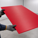 Red PE500 sheet held level in gloved hands, faint brushed grain across the face, pale workshop behind