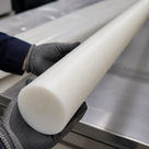Grey-gloved hands supporting a long white PET rod over a stainless bench, rounded end nearest camera