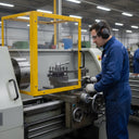 Machinist in blue overalls at a lathe behind a yellow-framed clear guard panel