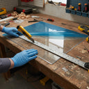 Gloved hands scoring a blue-film-covered clear sheet with a snap knife and steel rule on a bench