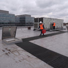 Black open-lattice matting running from an open roof hatch to rooftop air-handling plant, engineers in hi-vis
