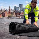 Roofer in hi-vis unrolling a roll of black open-lattice PVC walkway matting onto a flat membrane roof, city skyline behind