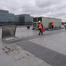 Light grey open-lattice matting from an open roof hatch to rooftop air-handling units, two engineers in hi-vis