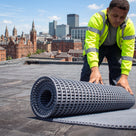 Roofer in hi-vis unrolling grey open-lattice PVC walkway matting on a flat roof, city skyline behind