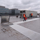 White open-lattice matting laid from an open roof hatch towards rooftop air-handling units, two engineers on site