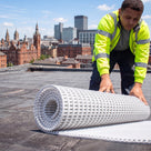 Roofer in hi-vis unrolling a roll of white open-lattice PVC walkway matting onto a flat membrane roof, city skyline behind