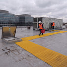 Yellow open-lattice matting laid from an open roof hatch out to rooftop air-handling plant, engineer with a toolbox