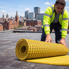 Roofer in hi-vis unrolling a roll of yellow open-lattice PVC walkway matting onto a flat membrane roof, city skyline behind