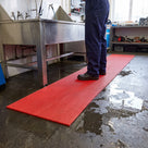 Red 15mm ribbed PVC drainage matting laid in front of a stainless-steel parts-washing sink in an engineering workshop, wet concrete floor beside it