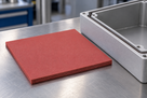 Dull red silicone sponge square resting on a brushed steel bench, open alloy enclosure at the right edge
