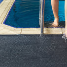 Close view of charcoal grey ribbed barefoot matting at a pool edge, a swimmer stepping out of the water onto it beside the ladder rail