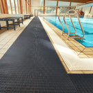 Charcoal grey ribbed barefoot matting laid the length of a tiled indoor swimming pool surround beside a chrome pool ladder