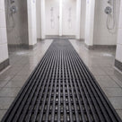 Charcoal grey ribbed barefoot matting running between two rows of showers in a communal shower block, water draining through the ribs