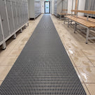 Dark grey ribbed barefoot matting runner down a changing room aisle between lockers and timber benches, wet footprints on the tiles