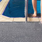 Close view of dark grey ribbed barefoot matting at a pool edge, a swimmer stepping out of the water onto it beside the ladder rail