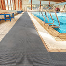Dark grey ribbed barefoot matting laid the length of a tiled indoor swimming pool surround beside a chrome pool ladder