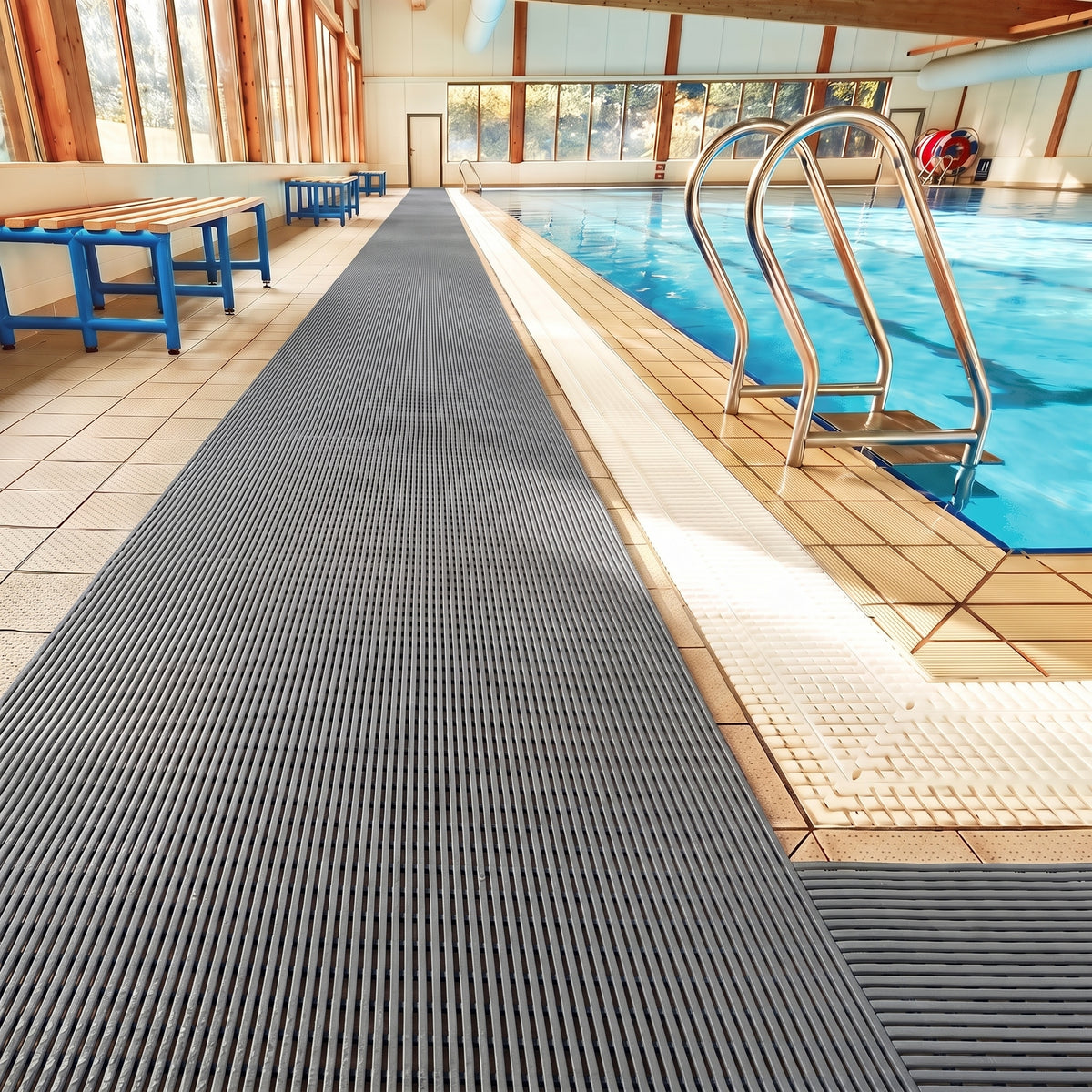 Dark grey ribbed barefoot matting laid the length of a tiled indoor swimming pool surround beside a chrome pool ladder