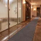 Dark grey ribbed barefoot matting as a wet-side walkway between a glass steam room and timber sauna door, drain channel alongside