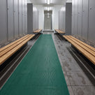 Green ribbed barefoot matting runner down a changing room aisle between lockers and timber benches, wet footprints on the tiles